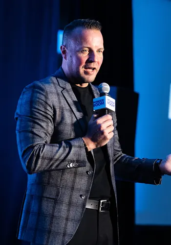 Speaker in a plaid blazer holding a microphone while addressing an audience on stage