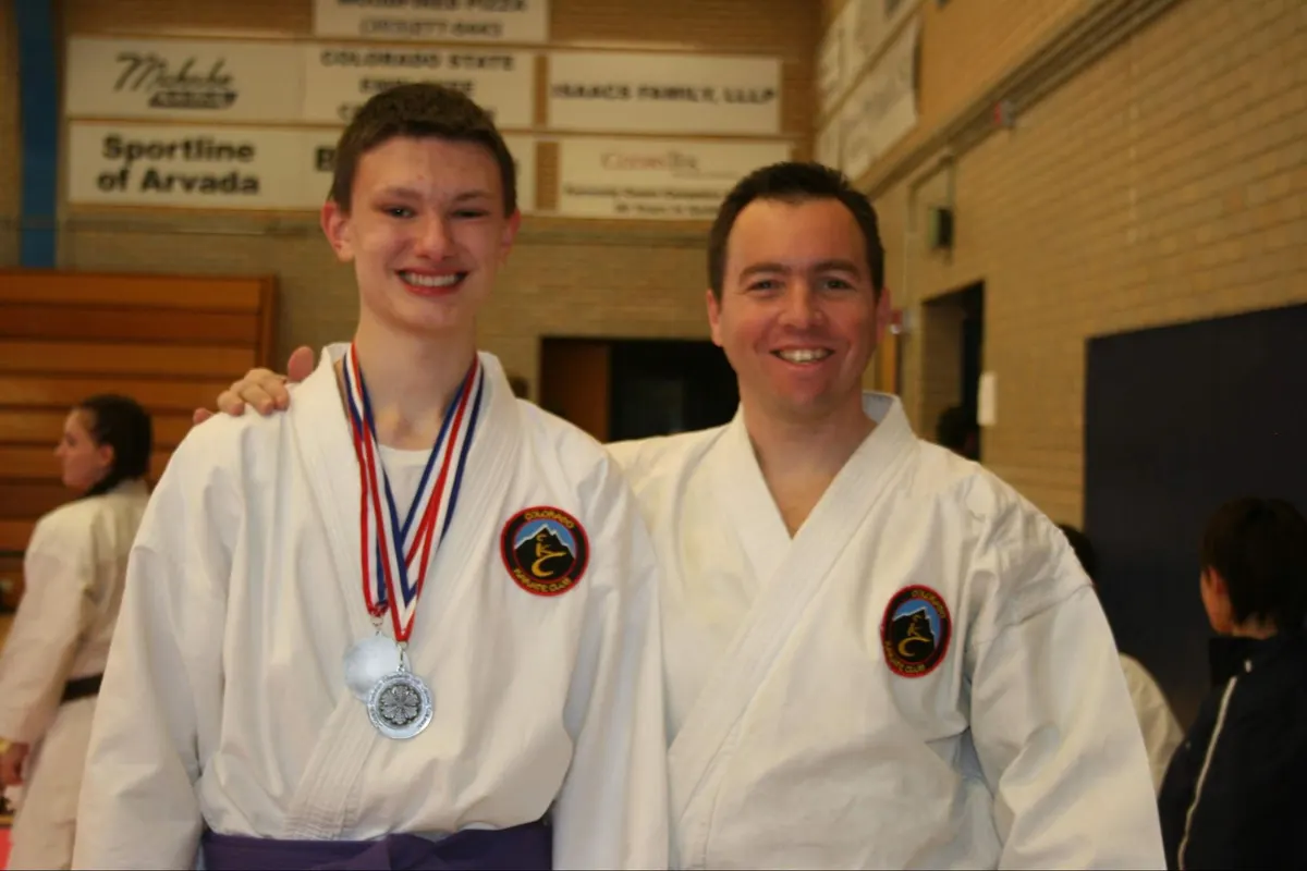 Teen karate student wearing a medal stands beside his smiling instructor at a tournament